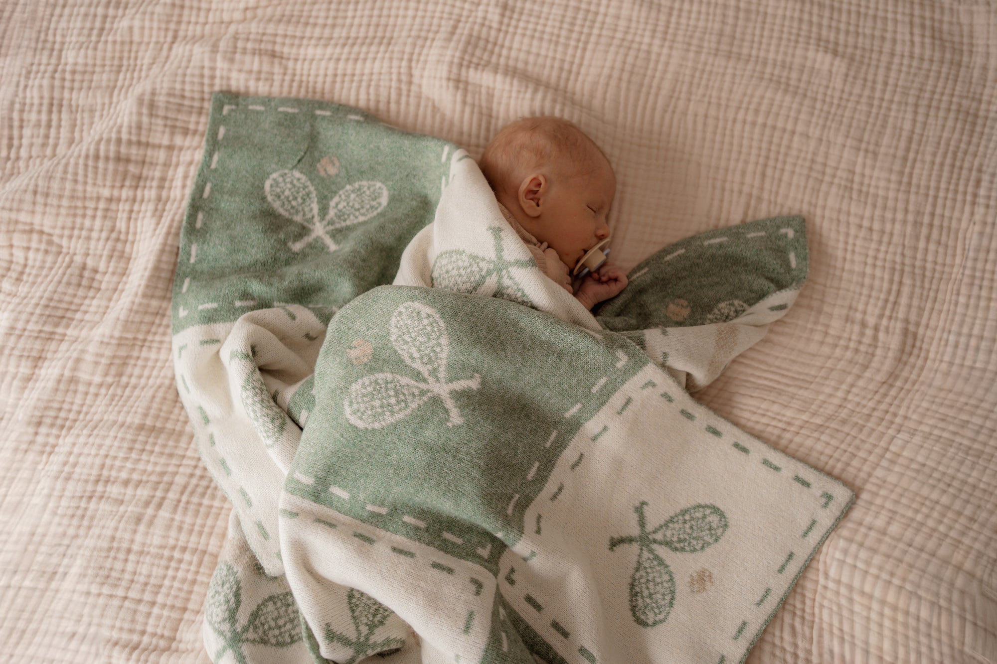 Baby wrapped in a green blanket with tennis patterns on a checkered bed.