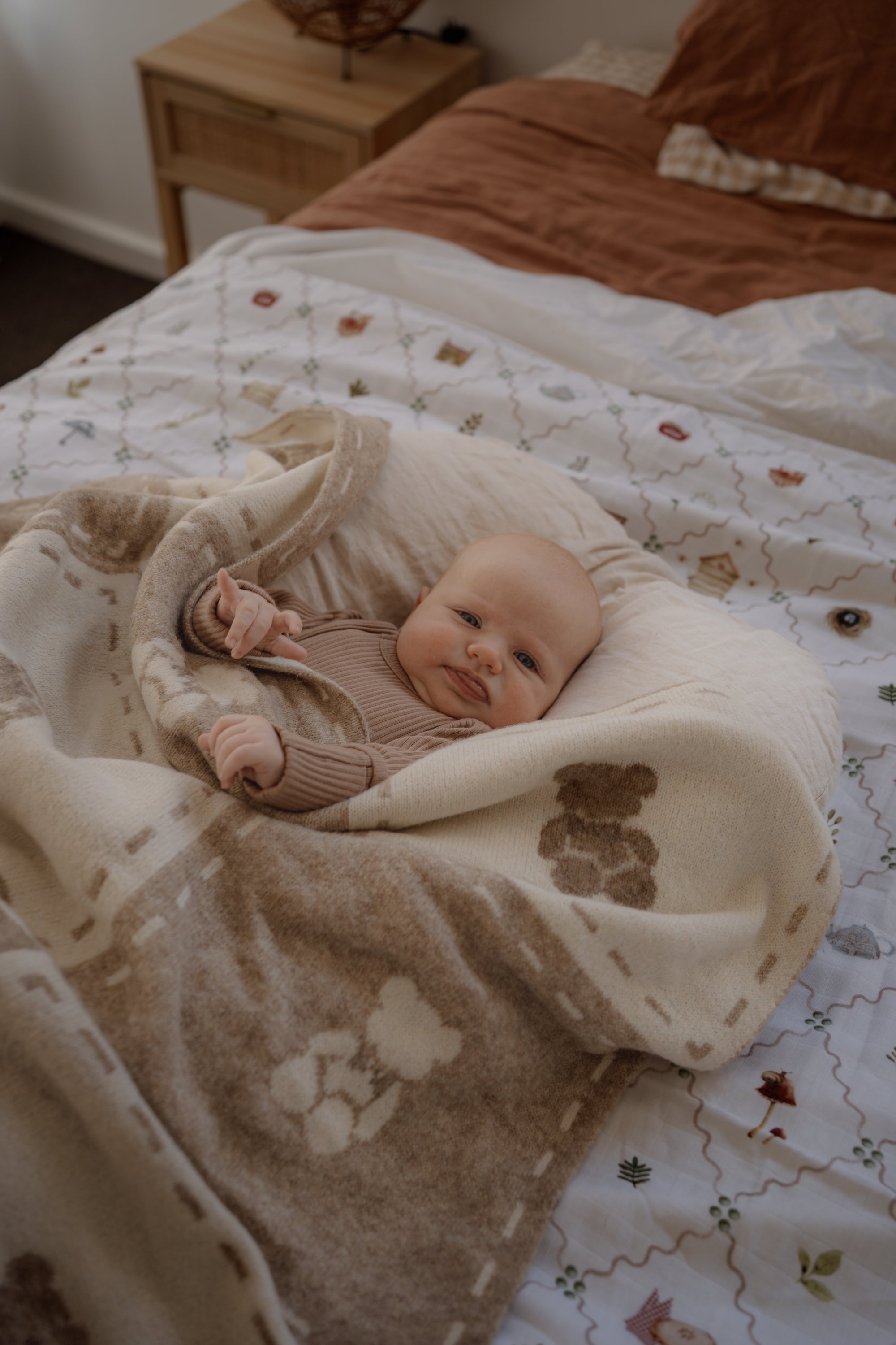 Baby wrapped in a blanket on a bed with a wooden nightstand in the background